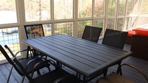 Eine überdachte Terrasse mit Holztisch und -stühlen, großen Fenstern und Blick auf ein Gewässer.