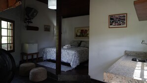 A bedroom with a bed, a small bedside table, a lamp, a painting on the wall, and a bathroom with a sink and countertop. - Fazenda Pedra Riscada (Nova Friburgo)