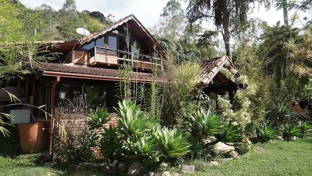A rustic wooden house with a balcony, surrounded by lush greenery and rocks. - Fazenda Pedra Riscada (Nova Friburgo)