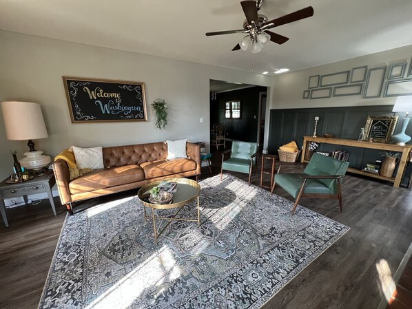Un salon avec un canapé en cuir marron, un tapis à motifs, une table basse et un ventilateur de plafond.