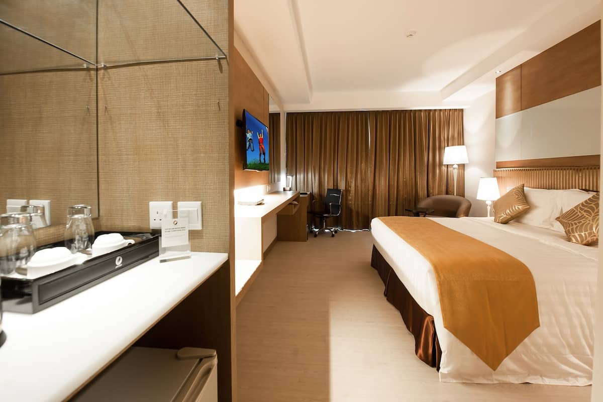 Ein Hotelzimmer mit einem großen Bett, einem Schreibtisch mit Computer, einem Sessel, einem an der Wand befestigten Fernseher und einem Spiegel.