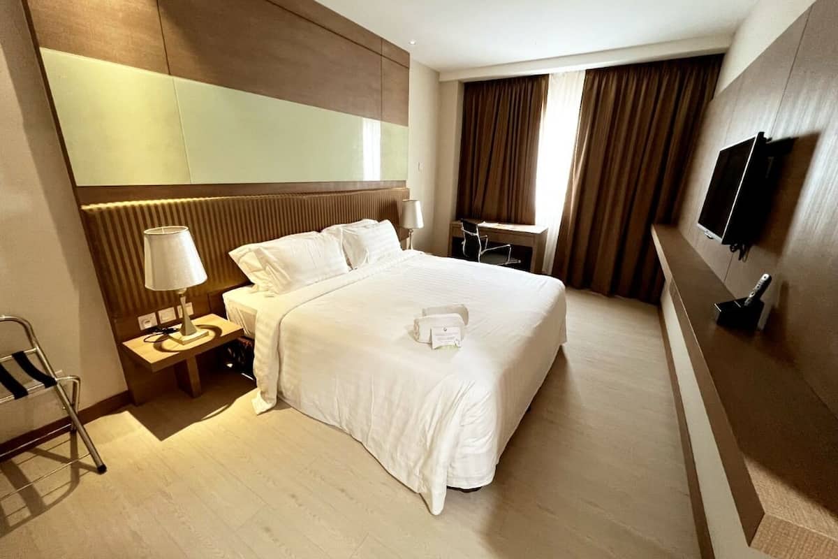 Ein Hotelzimmer mit einem großen Bett, einem Nachttisch mit Lampe, einem Sessel, einem Schreibtisch mit Computer und einem an der Wand befestigten Fernseher.