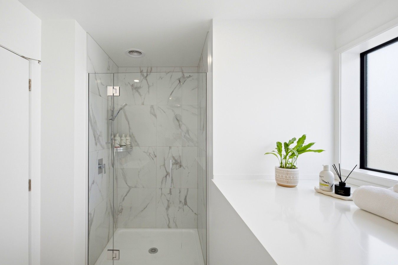 Un baño moderno con cabina de ducha de vidrio, paredes de mármol, encimera blanca y ventana.