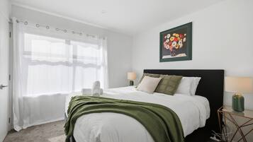 Una habitación de hotel con cama, mesita de noche, lámpara, un cuadro y ventana con cortinas.