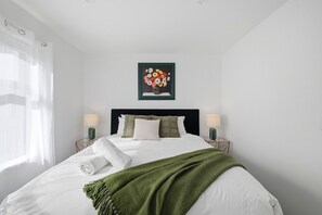 Un lit bien fait, avec du linge de lit blanc et un plaid vert, deux lampes de chevet et une œuvre d’art florale encadrée au-dessus du lit.