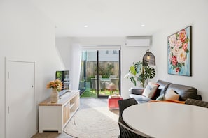 A modern living room with a sofa, a dining table, and a TV. There is a sliding glass door leading to a garden.