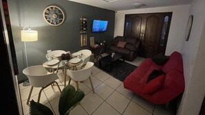 Ein Wohnzimmer mit Esstisch für vier Personen, einer Couch, einem roten Sessel und einem Fernseher an der Wand.