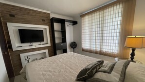 Ein Schlafzimmer mit Bett, Wandfernseher, Nachttisch, Lampe und einem Fenster mit Jalousien.