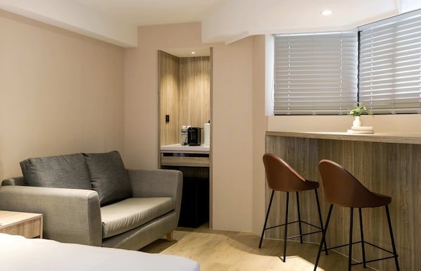 A modern room with a sofa, two bar stools, a small table, a window with blinds, and a plant on the windowsill. - Spring's Rhapsody Hotel (Taichung)