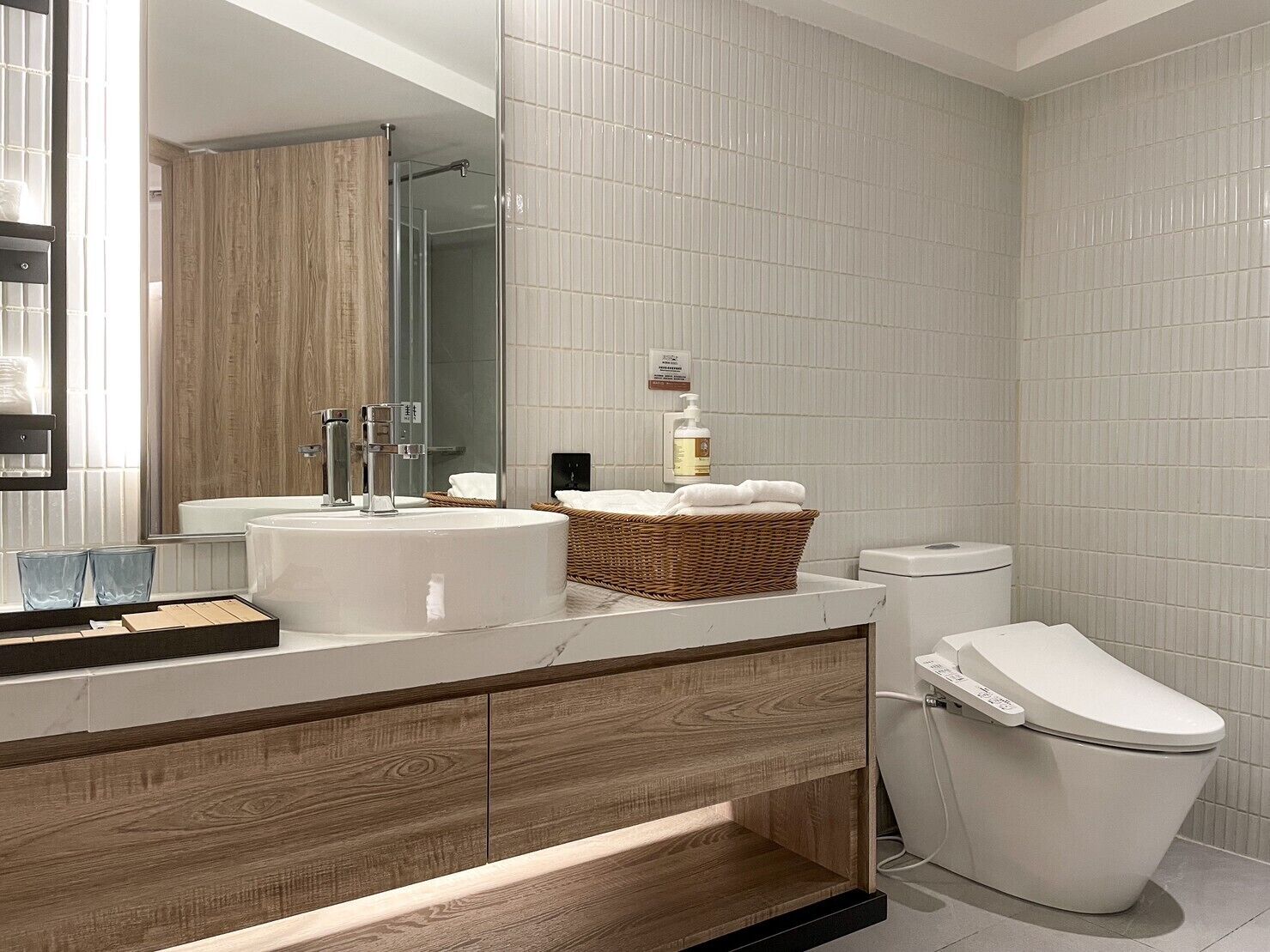 Een moderne badkamer met een houten wastafelmeubel, een grote spiegel, een witte wasbak, een toilet en een douchecabine met glazen wanden.