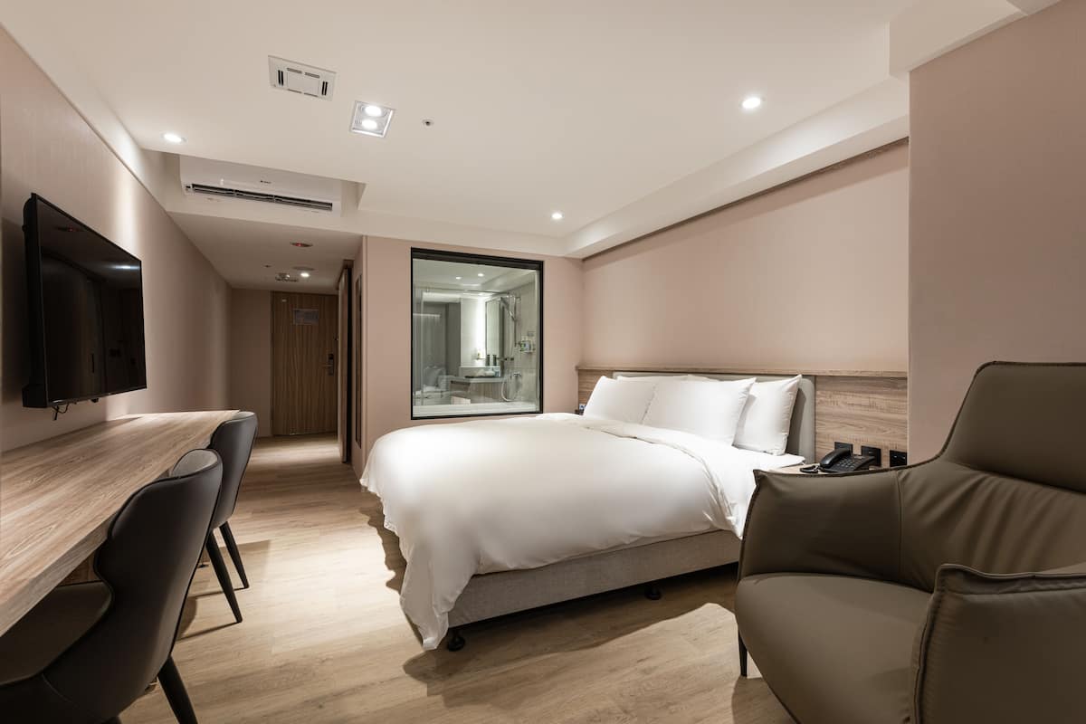 Ein modernes Hotelzimmer mit einem Holztisch, einem Bett mit weißen Leinen, einem braunen Ledersessel und einem großen Spiegel.