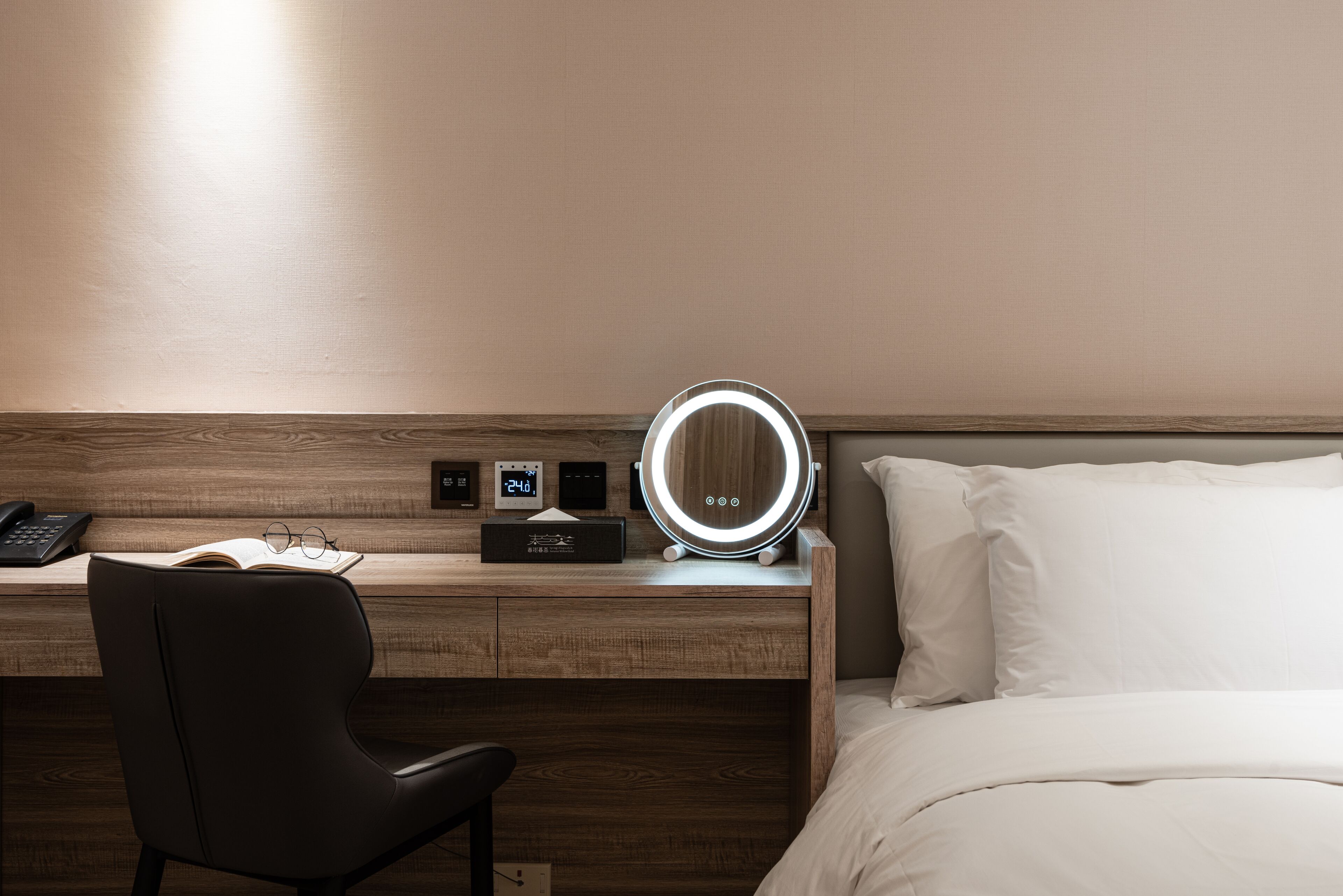 A hotel room with a wooden desk, a black chair, a bedside table with a mirror, a clock, and a bed with white linens.