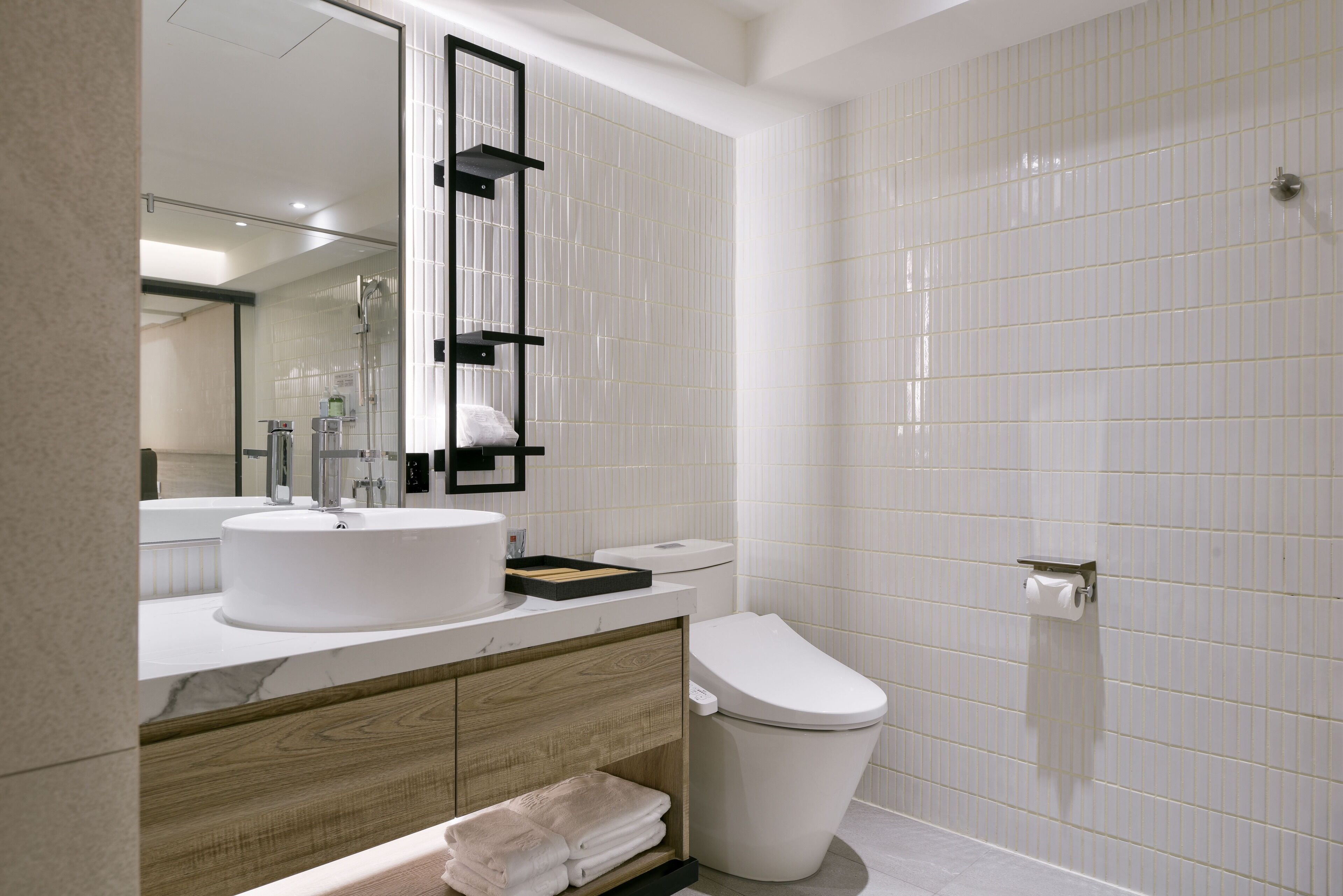 Ein modernes Badezimmer mit weißem Waschbecken, WC und Dusche. Es bietet einen Holztisch, einen großen Spiegel und geflieste Wände.