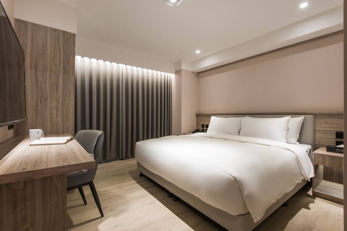 Ein modernes Hotelzimmer mit Schreibtisch aus Holz, einem bequemen Sessel, einem großen Bett mit weißer Bettwäsche und einem am Wand montierten Fernseher.