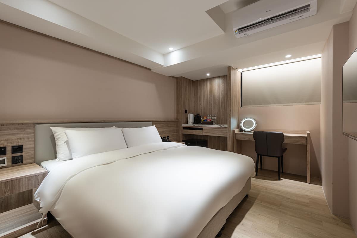 Ein modernes Hotelzimmer mit einem großen Bett, einem Schreibtisch mit Stuhl, einem Schminktisch mit Spiegel und einer Klimaanlage.