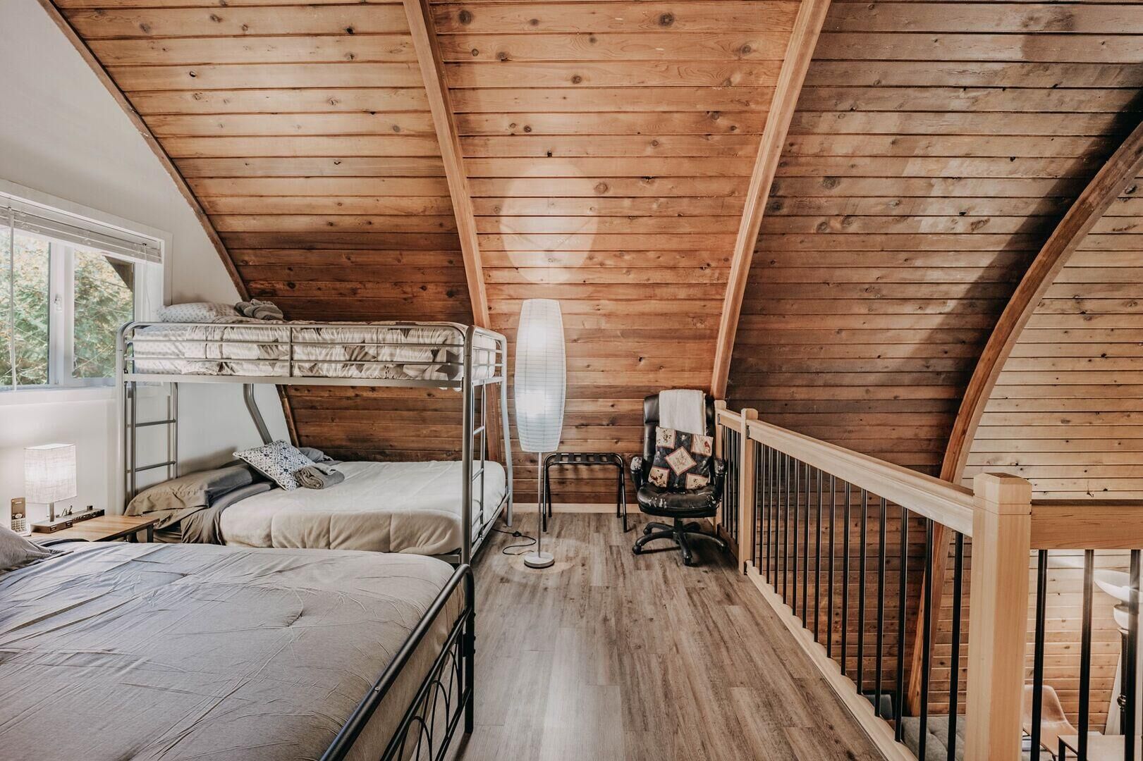 A cozy attic bedroom with a bunk bed, a chair, a standing fan, and a window with a view of trees.