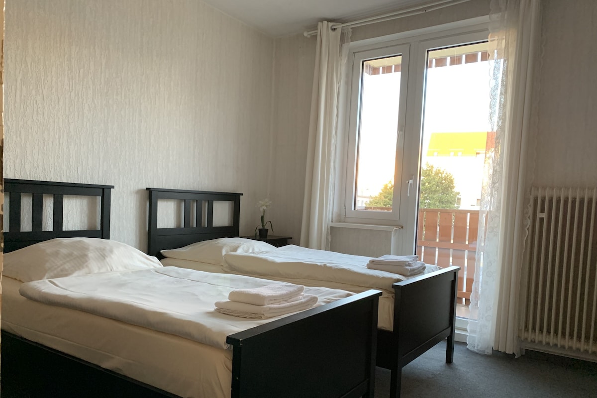 A hotel room with two single beds, a small table, and a window with curtains.