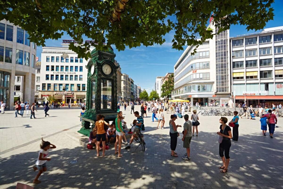 Ein belebter Marktplatz mit einem markanten Glockenturm, umgeben von modernen Gebäuden und Fußgängern.