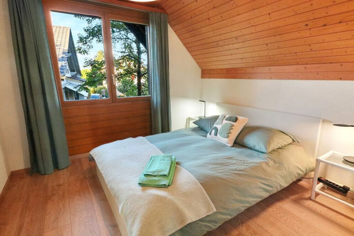 Une chambre avec un plafond en bois, une grande fenêtre avec des rideaux, un lit avec un drapé vert et des oreillers, et une table de chevet avec une lampe.