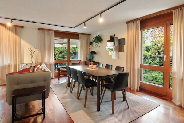 A dining area with a wooden table, black chairs, and a sofa. There are large windows with curtains, a plant on the windowsill, and a globe on the table.