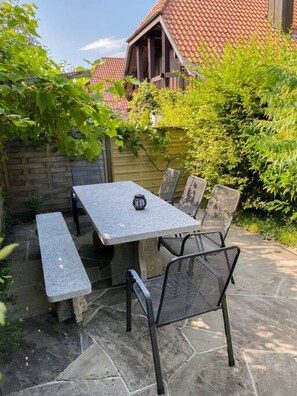 A patio with a stone table and metal chairs, surrounded by greenery and a wooden fence.