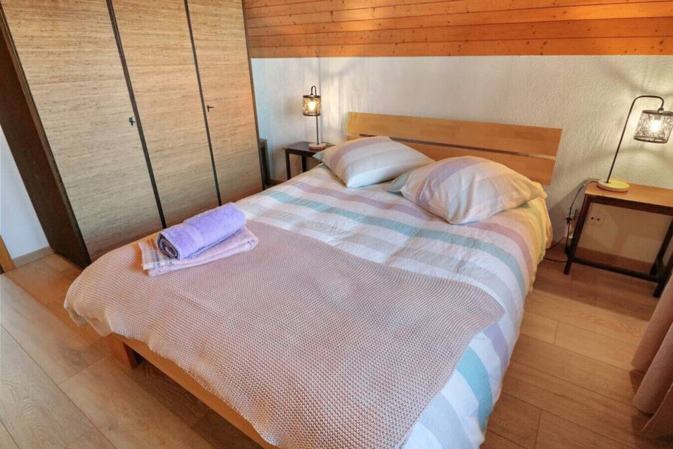 Una cama bien hecha con una manta a rayas, dos almohadas y una toalla doblada. Hay dos mesitas de noche con lámparas, un armario de madera y un cabecero de madera.