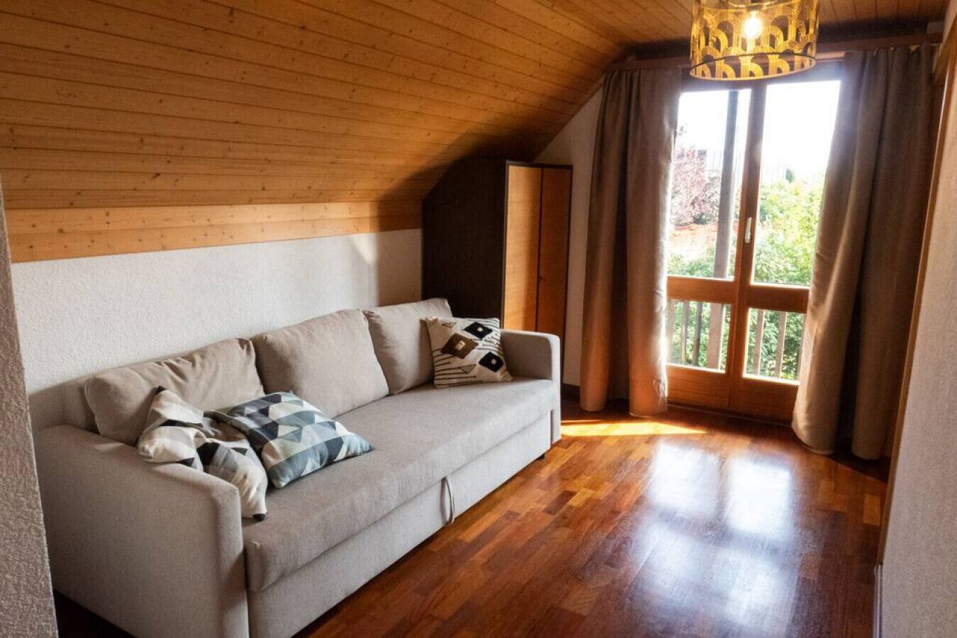 A cozy room with a wooden ceiling, a beige sofa with patterned cushions, and a large window with curtains.