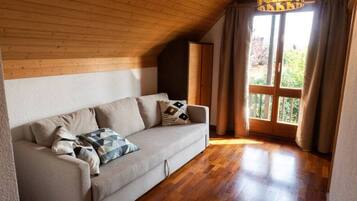 A cozy room with a wooden ceiling, a beige sofa with patterned cushions, and a large window with curtains.