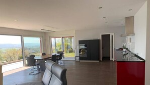 Ein großzügiger Wohnbereich mit einer großen Couch, einem Esstisch und Stühlen. Es gibt eine Küchenzeile mit einer roten Arbeitsplatte und Blick ins Freie durch große Fenster.