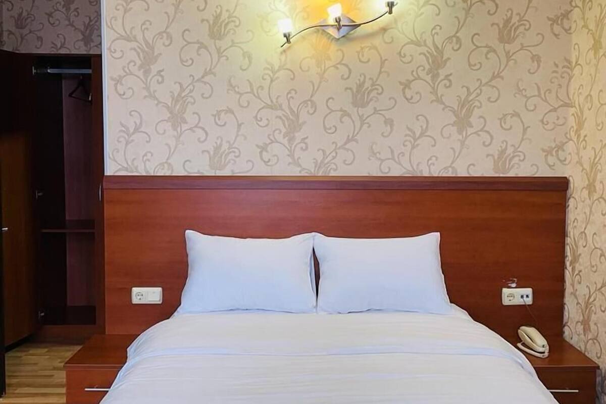 Una habitación de hotel con cabecera de madera, cama con ropa de cama blanca, un plaid verde, una lámpara de pared y un teléfono en la mesita de noche.