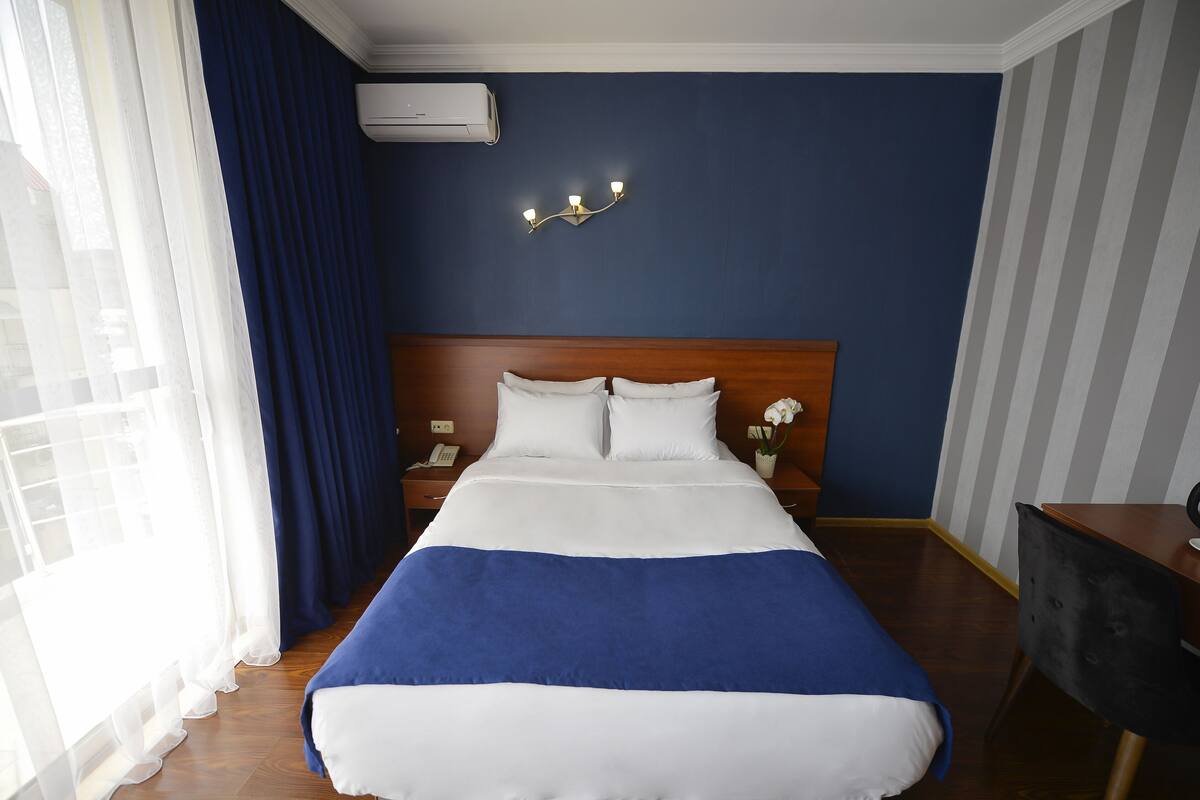 Una habitación de hotel con una cama grande, una mesita de noche, una lámpara y una ventana con cortinas.