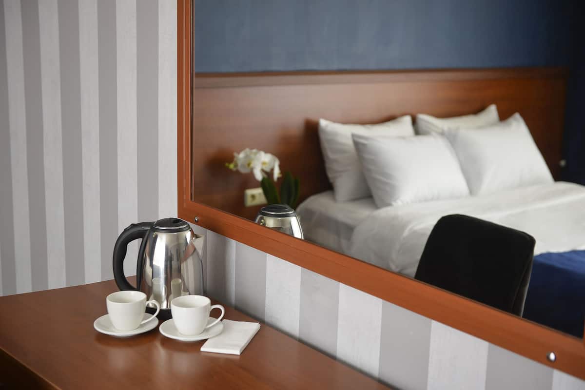 Habitación de hotel con escritorio de madera, dos tazas blancas, un hervidor eléctrico plateado y un espejo que refleja una cama con almohadas blancas y colcha azul.