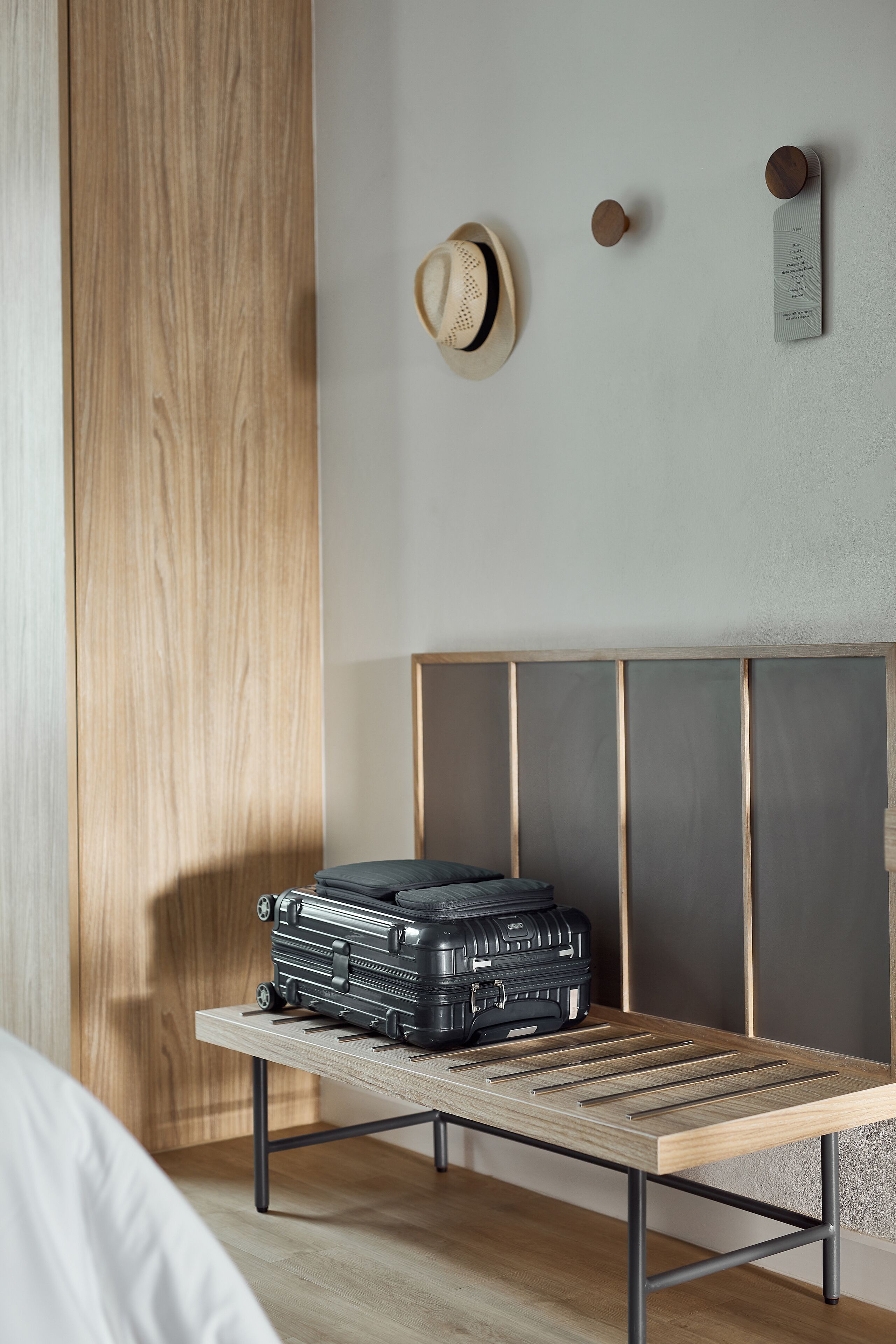 A neatly arranged room with a bed, a wooden nightstand, a suitcase, and a wall-mounted hat and clock.