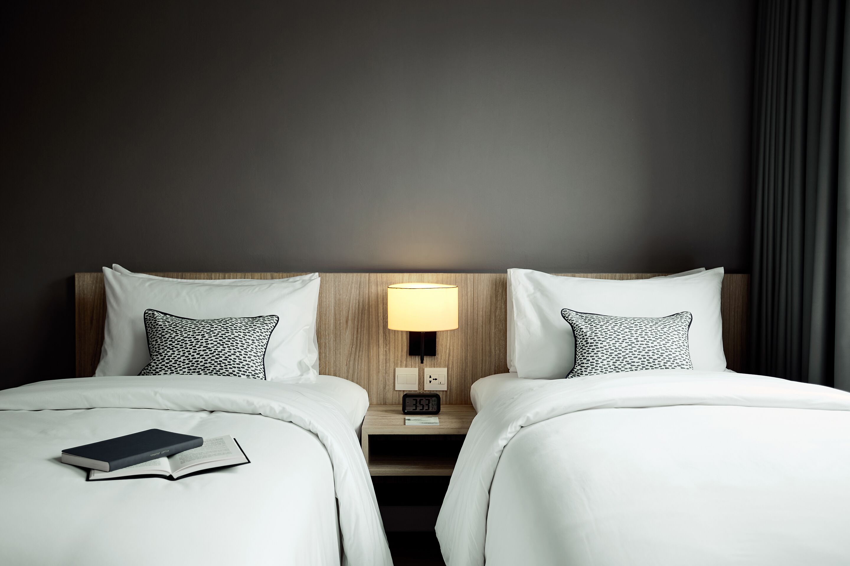 A hotel room with two beds, a bedside table with a lamp, and a digital clock.