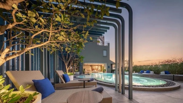 Zona de piscina exterior moderna con sillones y una mesa, rodeada de follaje y un edificio.