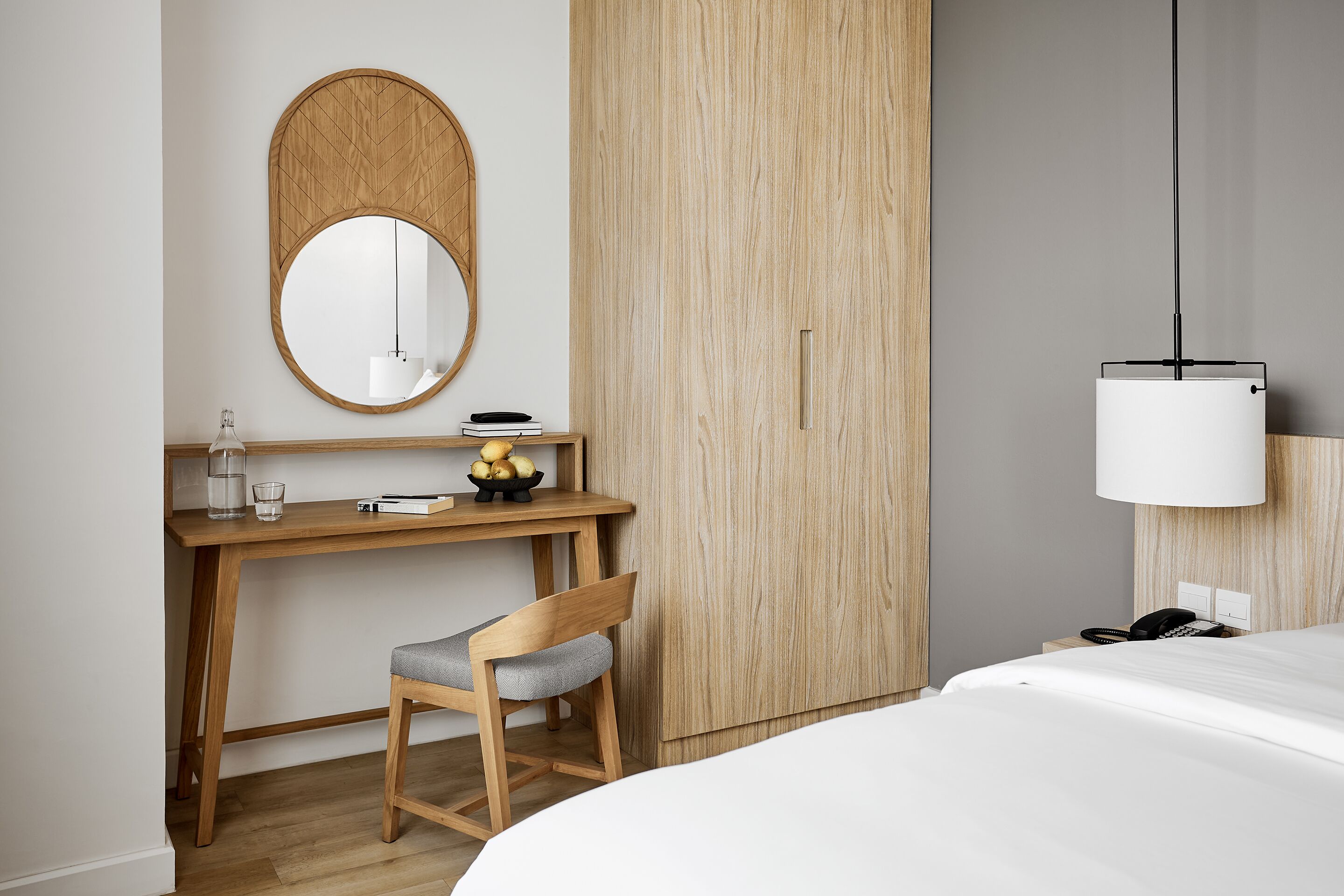 Une chambre d’hôtel moderne avec un bureau en bois, une chaise, un lit, un grand miroir et une lampe suspendue.