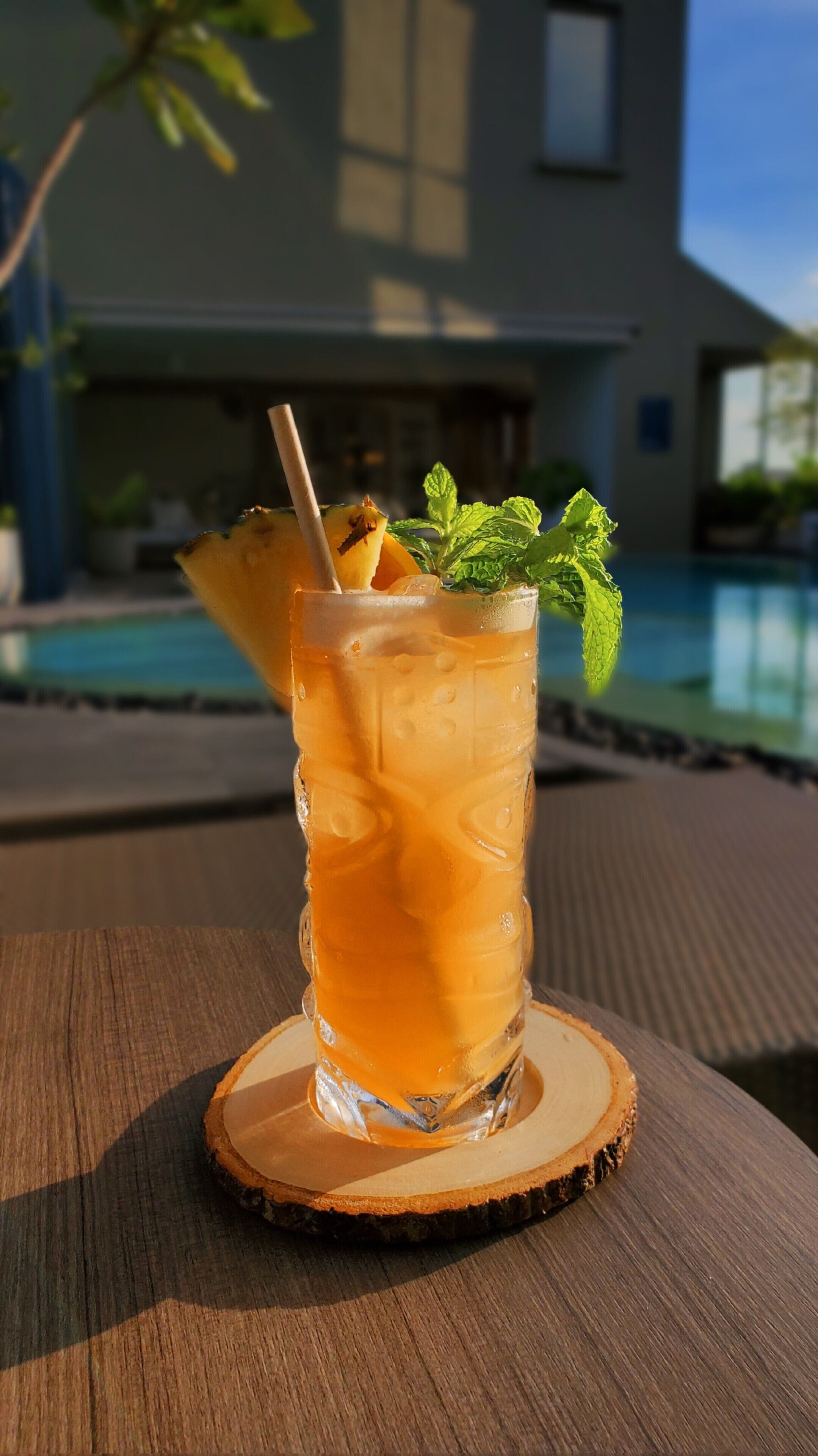 A glass of iced beverage with a straw and mint garnish on a wooden coaster by a poolside.