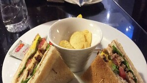 A plate with a sandwich cut in half, a bowl of chips, and a small packet of condiments. - Hotel SSV Grand (Hyderabad)