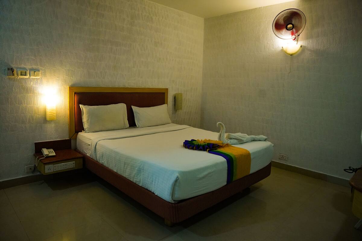 Quarto de hotel com cama, mesa de cabeceira, luminária e ventilador fixado na parede.
