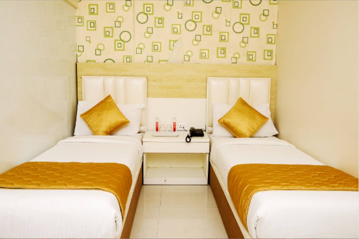 Quarto de hotel com duas camas, cada uma com cabeceira e colcha dourada. Um criado-mudo entre as camas possui uma luminária e alguns objetos. As paredes apresentam um padrão geométrico.