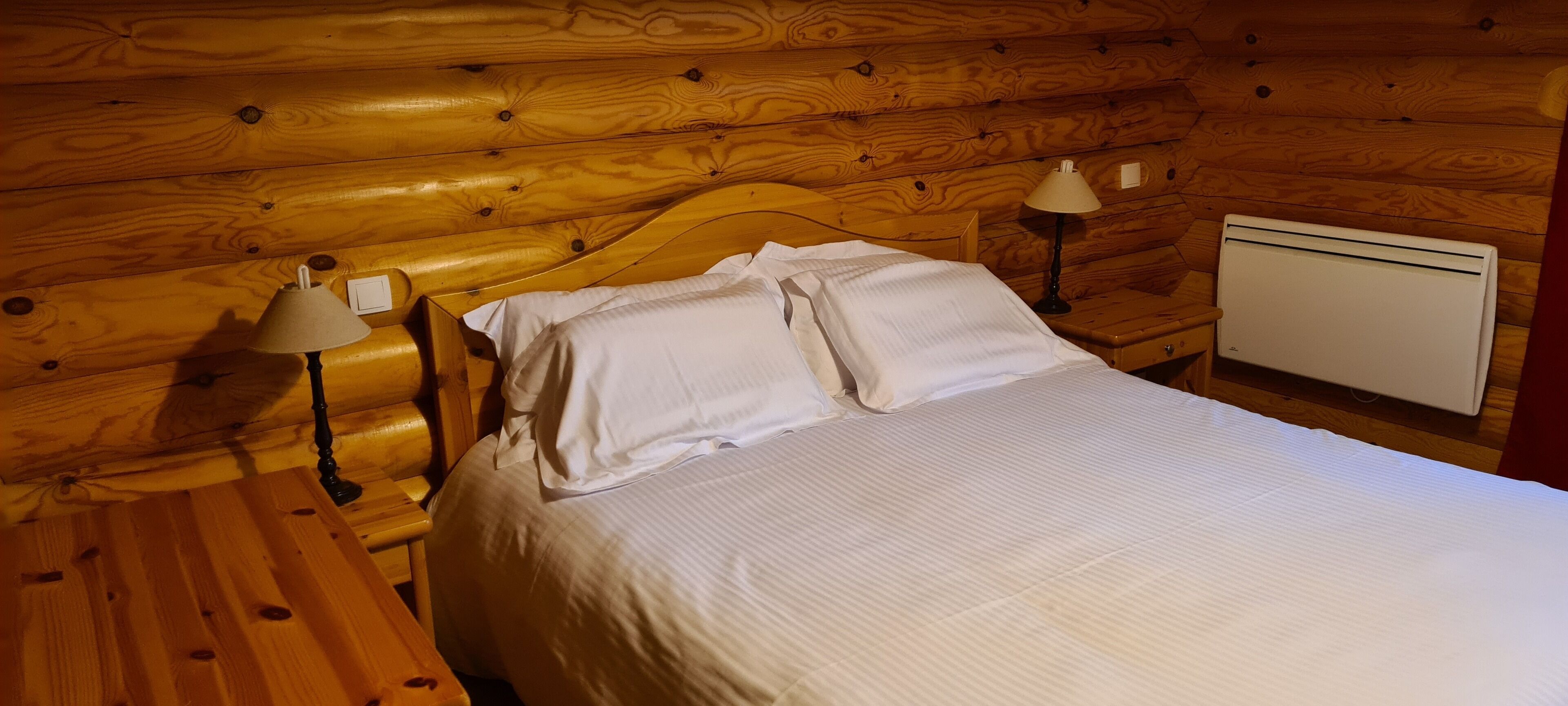 A wooden cabin room with a bed, two bedside lamps, and a heater.