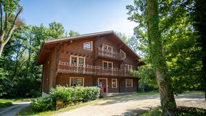 A two-story log cabin with a balcony, surrounded by trees and a gravel driveway. - Les Pins Sylvestres (Amneville)