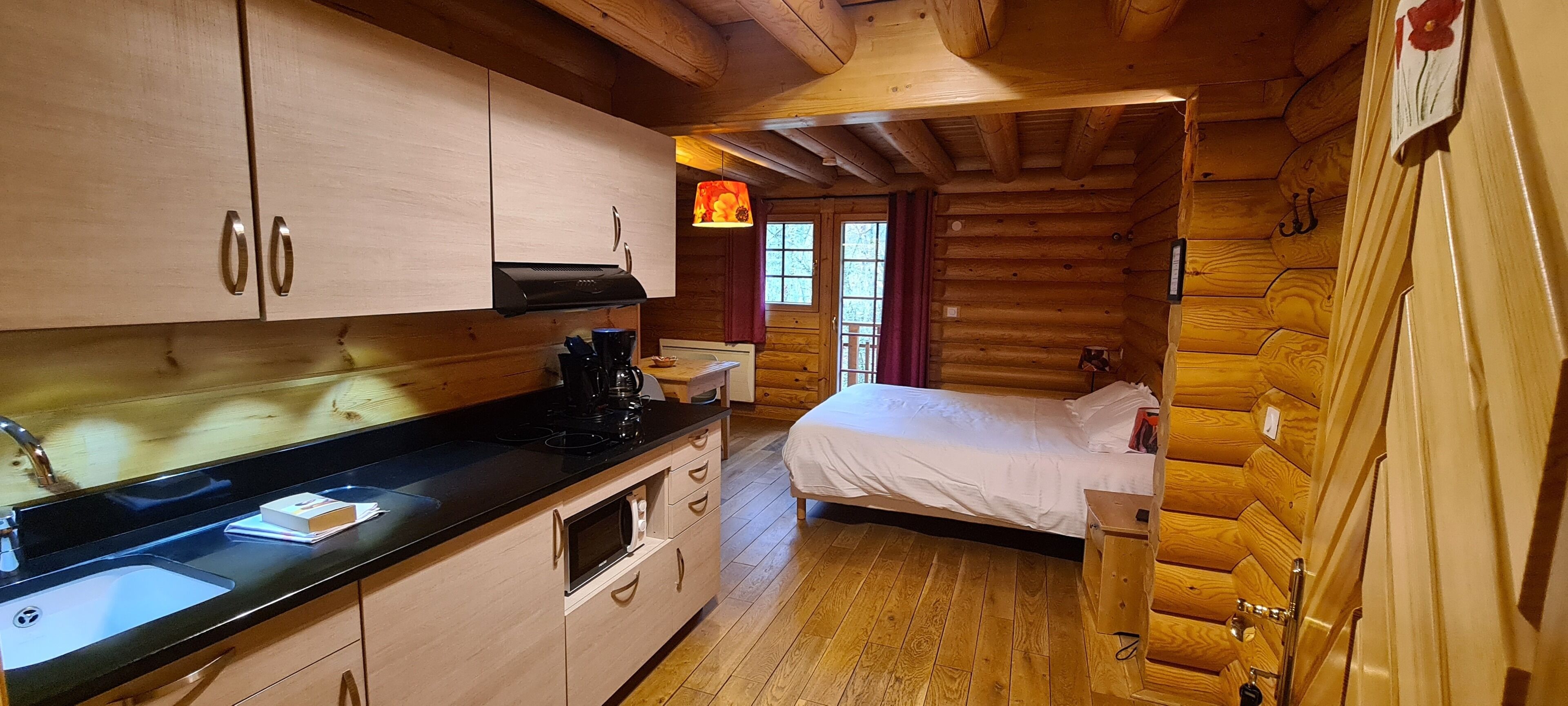 Uma cabana de toras aconchegante com cozinha equipada, incluindo fogão, micro-ondas e pia. Possui uma cama com edredom branco e uma janela com cortina vermelha.