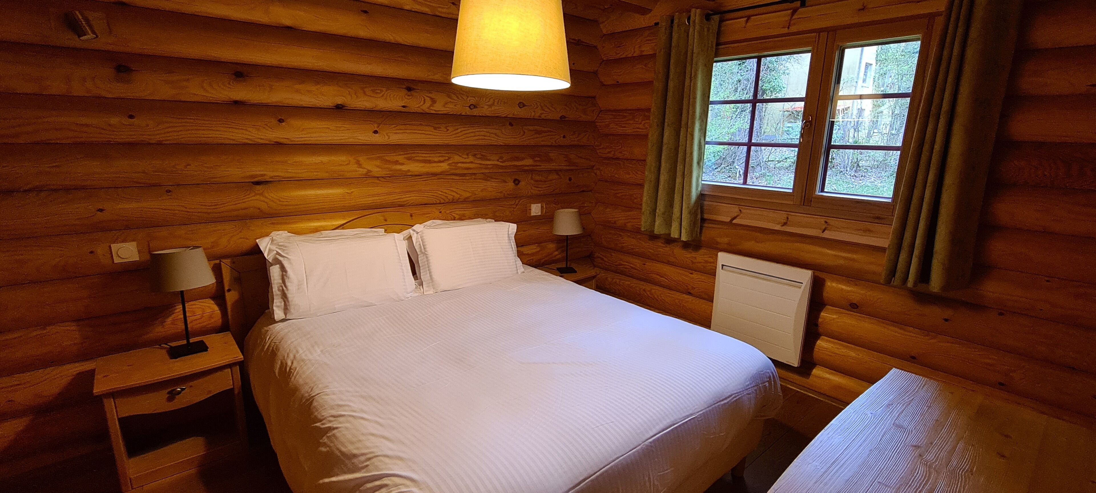 A wooden cabin bedroom with a bed, two bedside tables, a lamp, and a window with curtains.