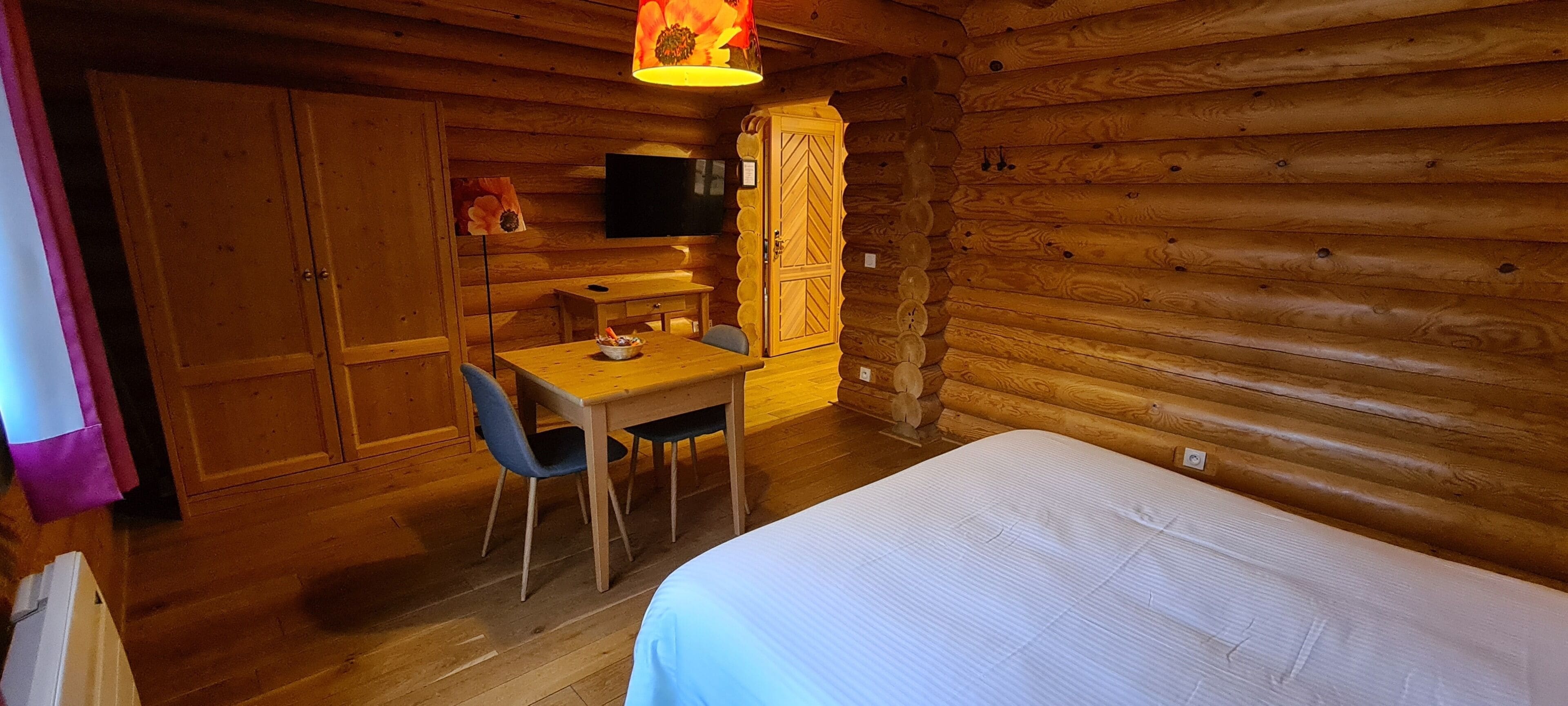 A wooden cabin room with a bed, a table with chairs, a TV mounted on the wall, and a wooden cabinet.