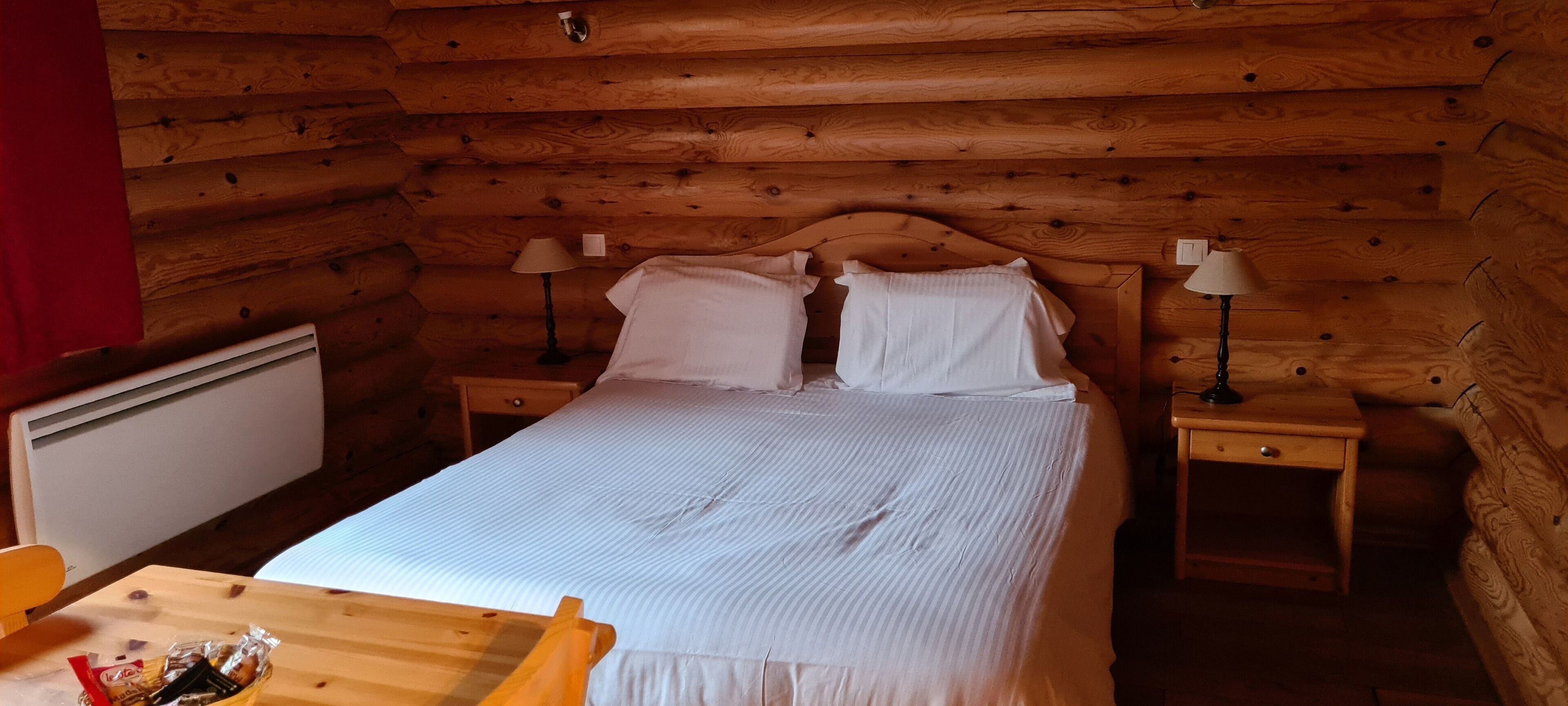 A wooden cabin room with a bed, two bedside tables, a lamp on each table, and a small table with a chair in the foreground.