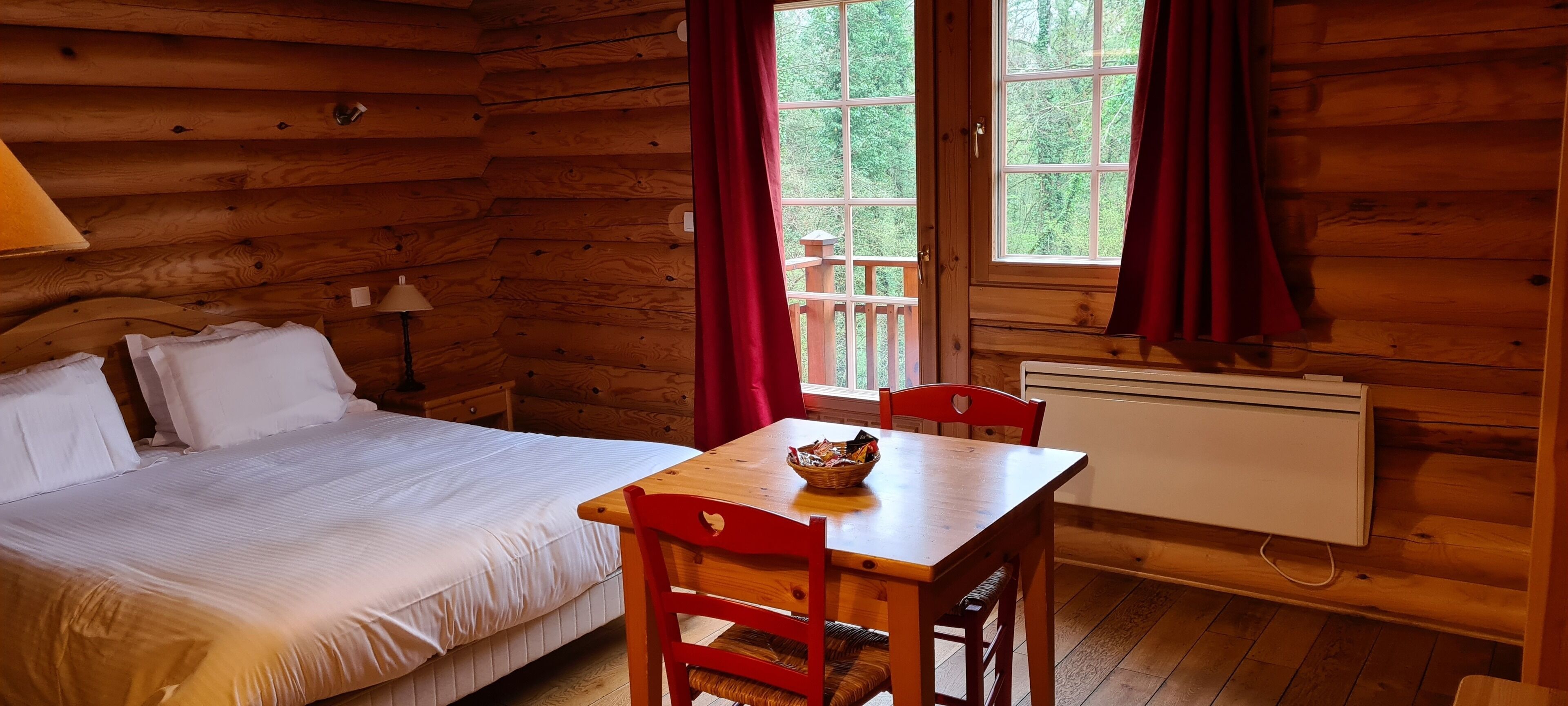 Une pièce de cabane en bois comprenant un lit, une table avec des chaises, une fenêtre avec des rideaux et un radiateur.