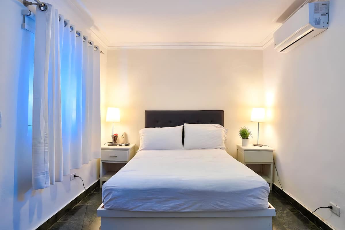 Un lit bien fait, avec du linge de lit blanc, deux tables de chevet avec des lampes et un climatiseur fixé au mur.