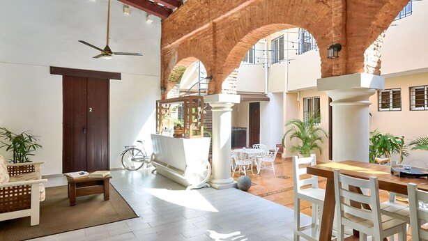 Een ruime binnenruimte met een hoge plafond en een plafondventilator, een houten tafel met stoelen. Er staan potplanten en staat een fiets op de achtergrond.
