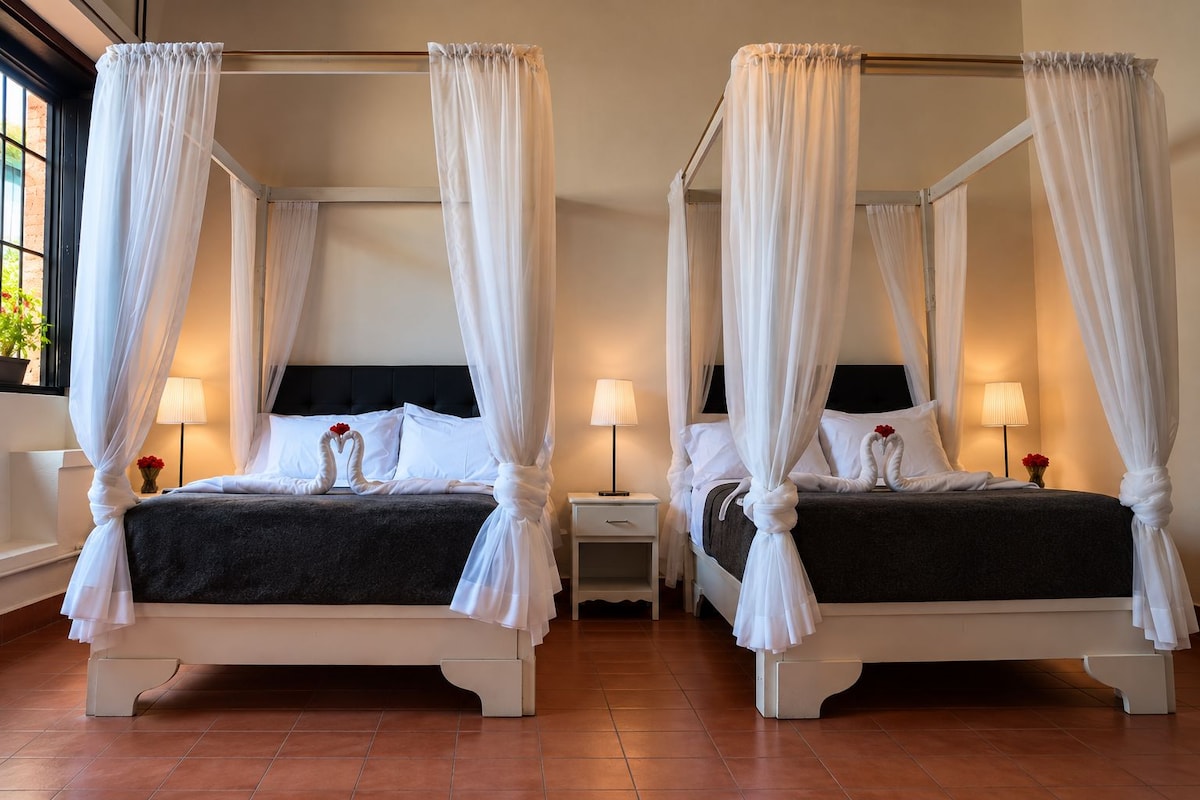 Two four-poster beds with white canopies, each with a bedside table and lamp, in a room with tiled flooring.